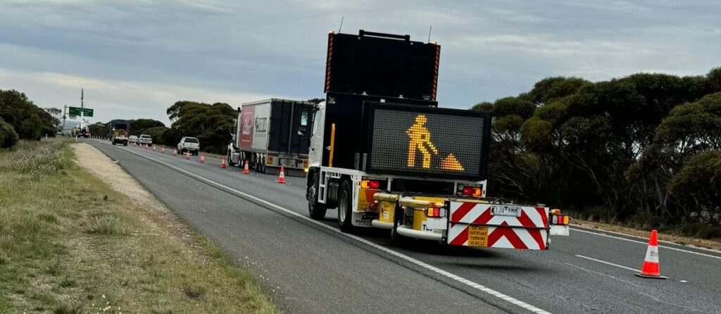 Traffic control vehicle with digital warning sign displaying construction worker symbol on highway, surrounded by traffic cones and vehicles, illustrating safety measures for road work.