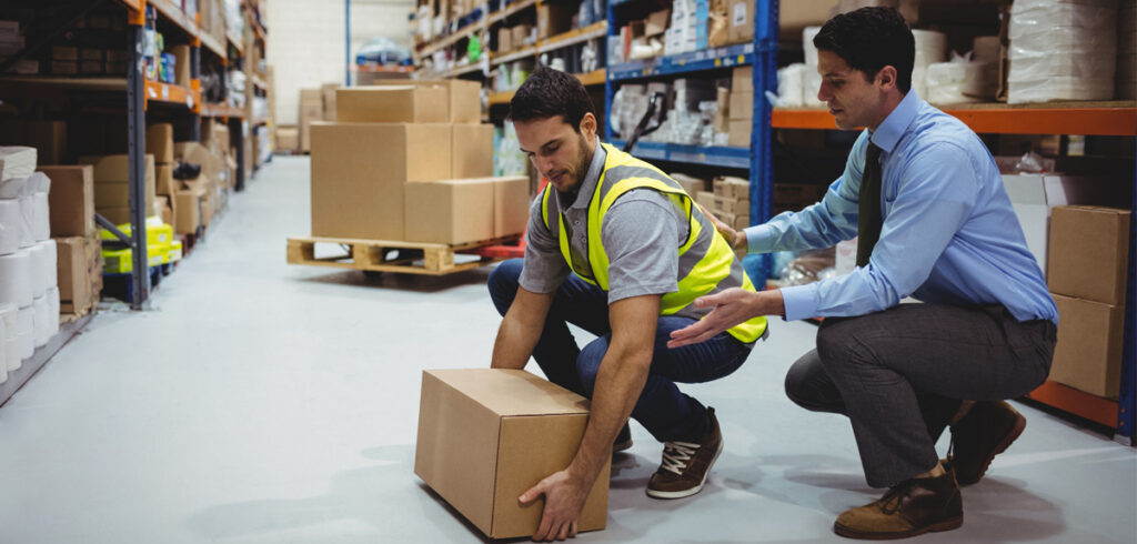 Worker in safety vest lifting box with guidance from supervisor in warehouse, emphasizing manual handling techniques for workplace safety.